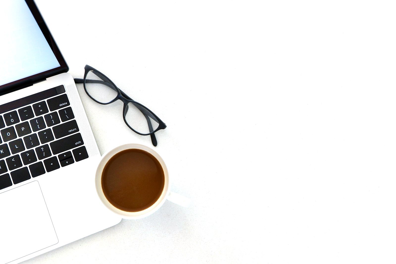Vue du dessus : un ordinateur portable, une paire de lunettes et une tasse de café sur un bureau blanc.