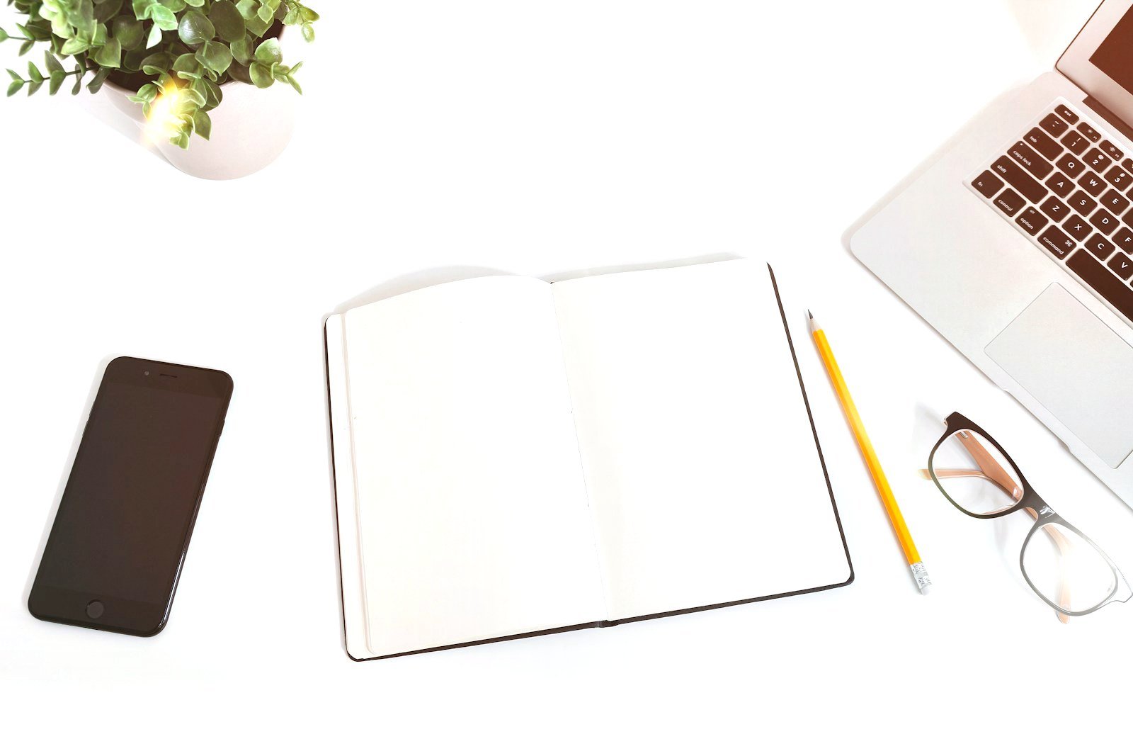 Vue du dessus : un carnet ouvert, un crayon, des lunettes, un téléphone et un ordinateur portable sur un bureau blanc.